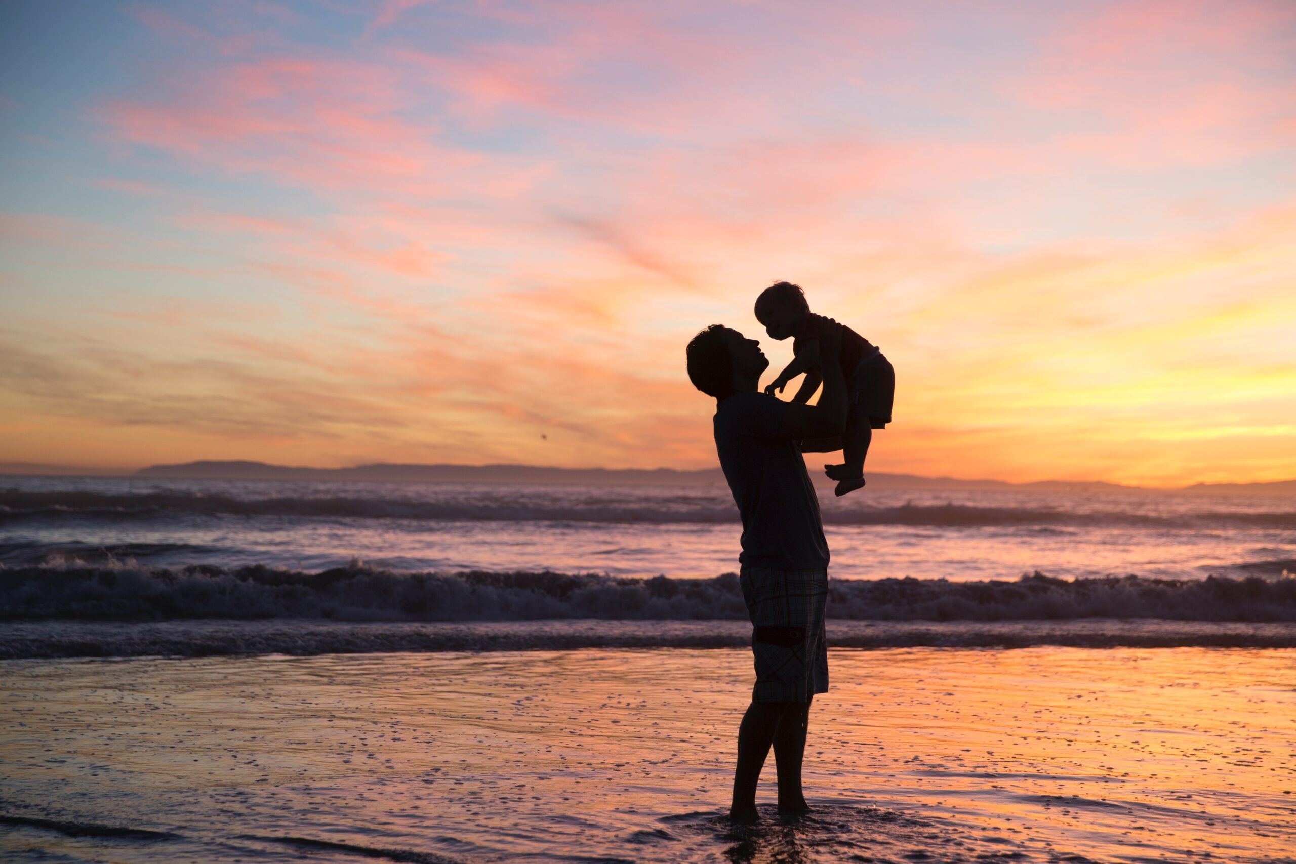A father holding a toddler on a beach at sunset