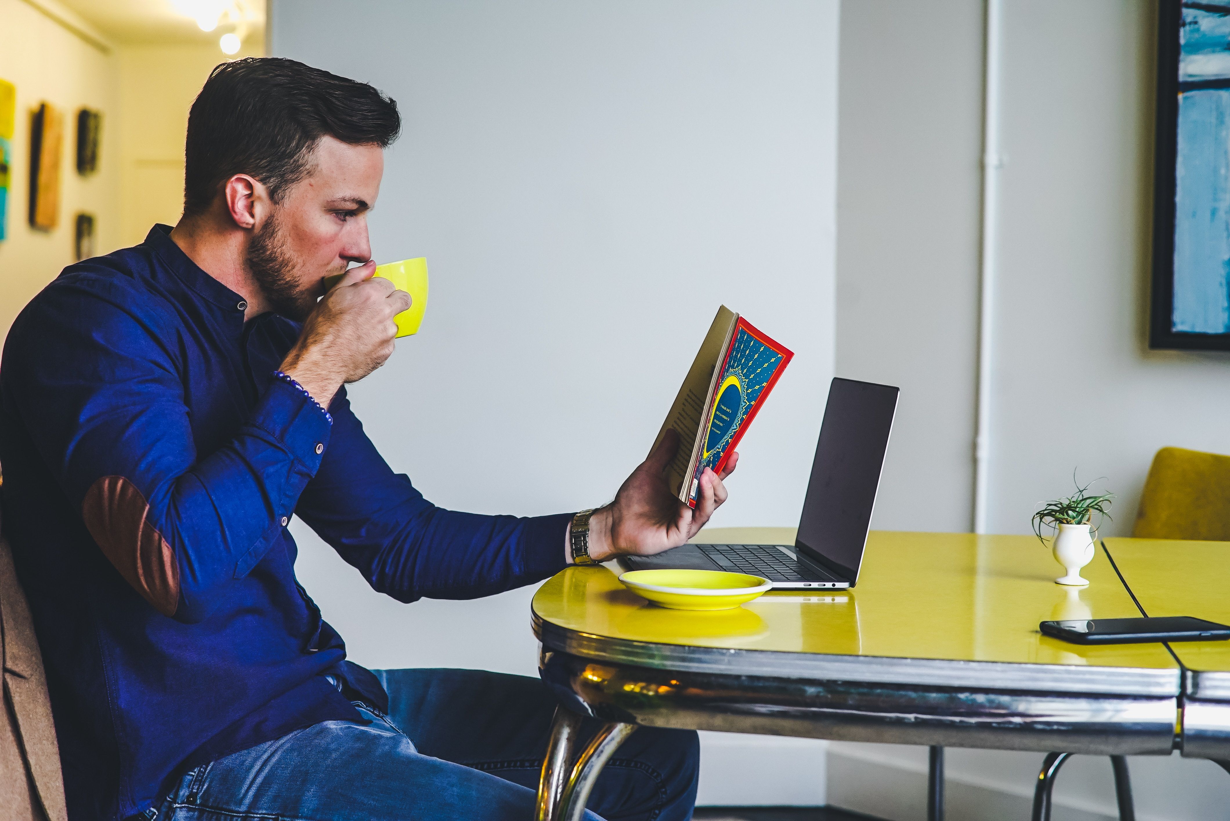 man reading a book at a yellow table with a yellow cup in his hand and a laptop open in front of him.
